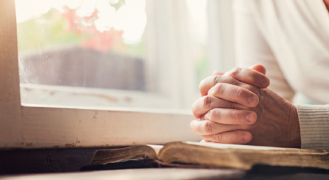 Folded hands praying over a Bible by a window