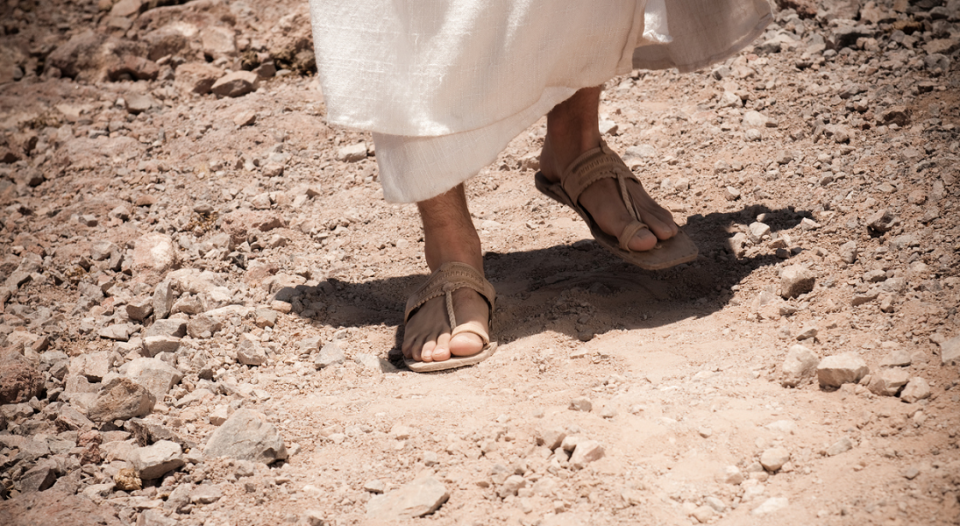 Feet beneath a white robe wearing sandles on a dusty pasth.