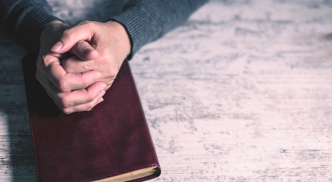 Two hands folded in prayer on a Bible