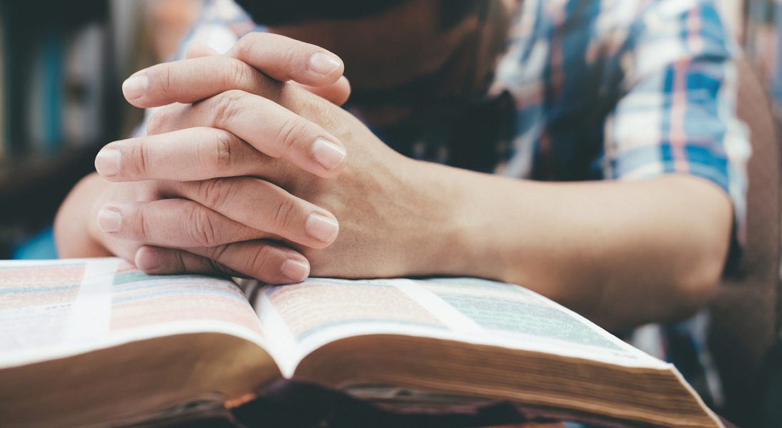 A closeup of a man's hands folded in prayer on an open Bible.