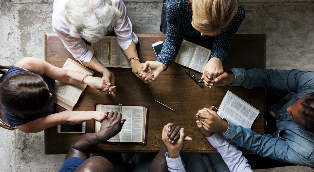 A group of people at a table hold hands in prayer.