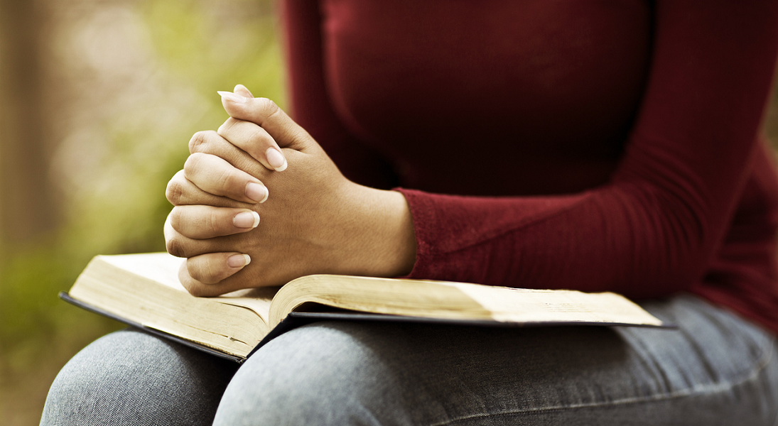 A woman's hnds folded in prayer resting on a open Bible on her lap.